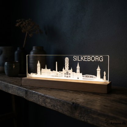 A LED night light with a laser engraved silhouette of Silkeborg, Denmark, glowing with warm light on a dark wooden base. The lamp is styled on a rustic wooden shelf against a dark, moody background with decorative vases.